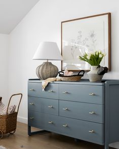 a blue dresser with flowers and a basket on top in a white room next to a lamp