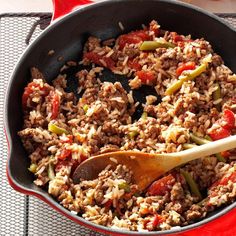 a skillet filled with rice and vegetables on top of a red table cloth next to a wooden spoon