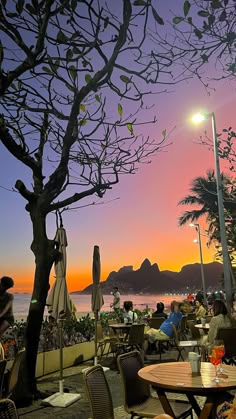 people are sitting at tables on the beach as the sun sets in front of some mountains