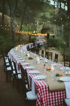 a long table is set up for an outdoor dinner party with lights strung from the trees