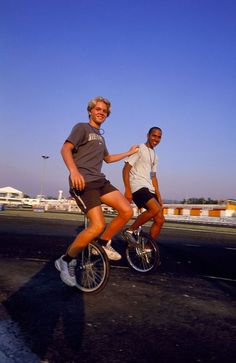 two men are riding bikes on the road in front of an airport terminal and one is smiling at the camera