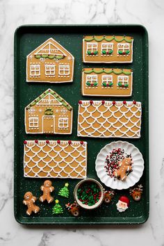 gingerbread houses are arranged on a green tray with white plates and small christmas decorations