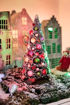 a small christmas tree on top of a snow covered ground next to buildings and a teddy bear