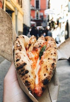 someone holding up a slice of pizza in the middle of an alleyway with people walking by