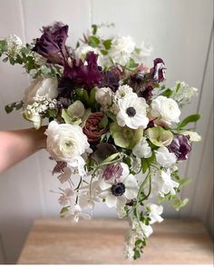 a bouquet of white and purple flowers in someone's hand on a wooden table