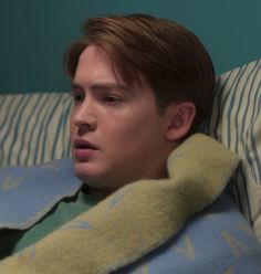 a young man laying in bed with a blanket around his neck and looking at the camera