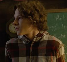 a young man standing in front of a chalkboard with writing on it and a clock behind him