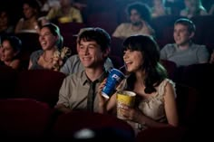 two people sitting next to each other in a movie theater holding popcorn cups and smiling