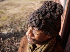 a man wearing glasses and a knitted hat looks off into the distance while leaning against a brick wall