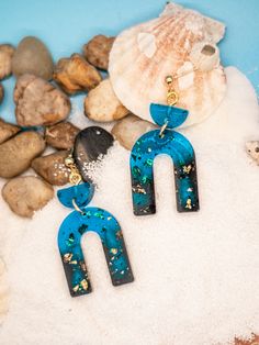 two pairs of blue and black earrings sitting on top of some rocks next to seashells