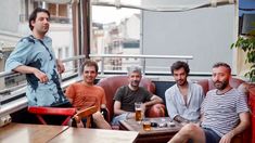 a group of men sitting around a table with beers in front of them and one man standing up