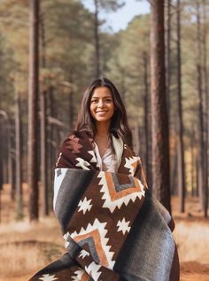 a woman standing in the woods with a blanket on her shoulders and smiling at the camera