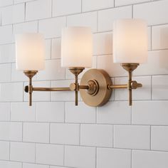 a bathroom light fixture with three lights on the wall and white tiles in the background