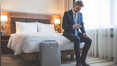 a man in a suit sitting on a bed looking at his cell phone while holding a suitcase