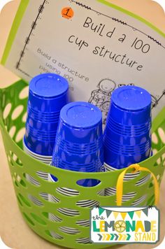 blue cups are in a green basket with a yellow ribbon around the edges and a sign that says build a 100 cup structure