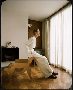 a man sitting on top of a wooden bench in front of a window with white curtains