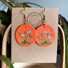 two orange earrings with flowers painted on them are sitting on a card next to a potted plant