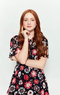 a woman with long red hair wearing a floral dress and holding her chin on her hand