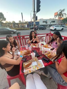 four women sitting at a red table eating hotdogs and sandwiches in front of them