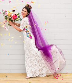 a woman in a white dress and purple veil holding a bouquet of flowers while standing next to a brick wall