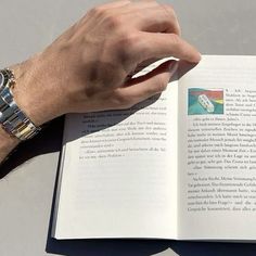 a person is holding an open book in their hand while sitting at a table with a watch on it