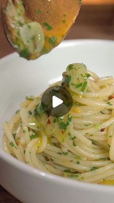 a spoon with some noodles and olives in it on a white bowl next to a wooden table