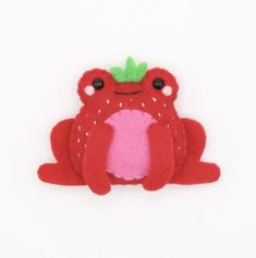 a red stuffed frog with a green leaf on its head and eyes sitting in front of a white background