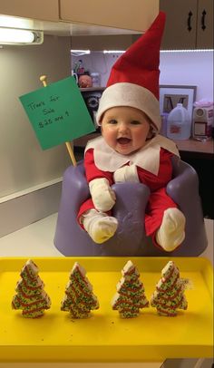 a baby dressed as an elf sitting in a chair with christmas trees on the table