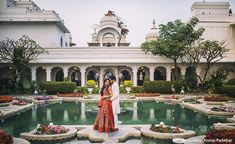 two people standing in front of a pool surrounded by flowers and greenery with an ornate building in the background