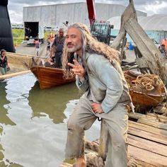 a man with long hair standing in the water next to a wooden boat and people