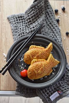 two fried fish on a black plate with chopsticks and cherry tomatoes next to it