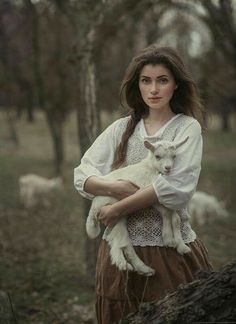 a woman holding a baby goat in her arms while standing next to a tree trunk