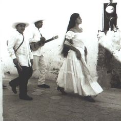 a woman in a white dress is walking with two men and a guitar on the street