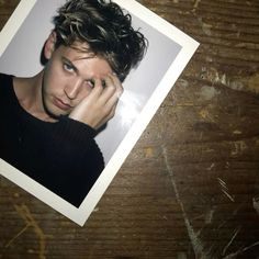 a polaroid photo of a young man with curly hair on a wooden table, holding his hand to his face