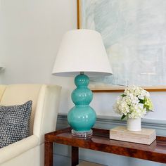 a blue lamp sitting on top of a wooden table next to a white chair and a painting