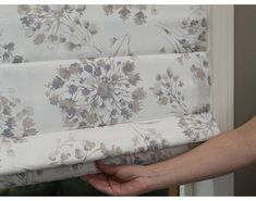 a hand is pulling the valance off of a flowered roman blind in front of a window