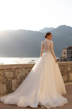 a woman in a wedding dress is standing on a stone wall by the water and mountains