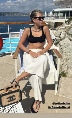 a woman sitting in a chair next to the ocean with a suitcase on her lap