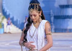 a woman in a white dress and gold jewelry is looking down at her cell phone