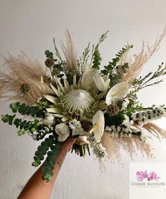 a hand holding a bouquet of white flowers and greenery in front of a wall