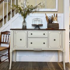 a white dresser sitting under a stair case next to a table with flowers on it