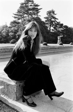 black and white photograph of woman sitting on stone wall next to water fountain with trees in background