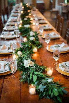 a long table with candles and greenery on the top is set for an elegant dinner