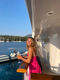 a woman in a pink dress holding a glass of wine on a boat near the water