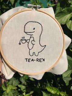 a person holding up a cross - stitch hoop with a drawing of a t - rex on it