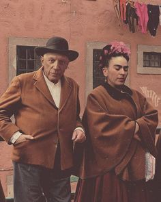 an old man and woman standing next to each other in front of a pink building