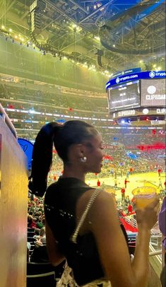 a woman standing in front of a crowd at a basketball game holding a drink and looking off into the distance