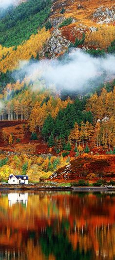 a house on the shore of a lake surrounded by trees with autumn foliage in the background