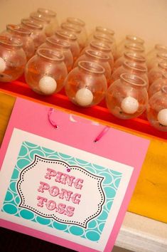 a pink and blue table topped with lots of small plastic cups filled with liquid next to a sign that says ping pong toss