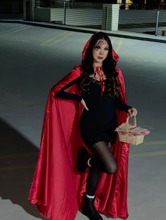 a woman dressed in a red cape and black dress holding a basket while standing on the street